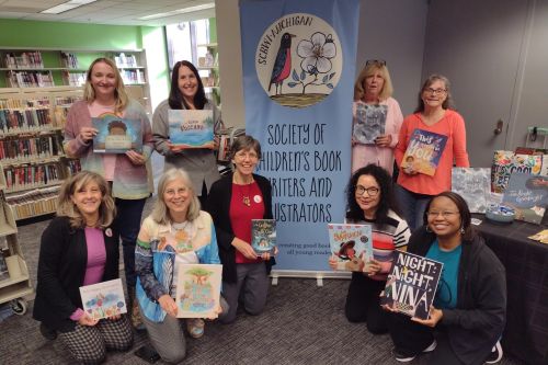 A group of nine authors with their books by the Society of Children's Book Writers and Illustrators banner
