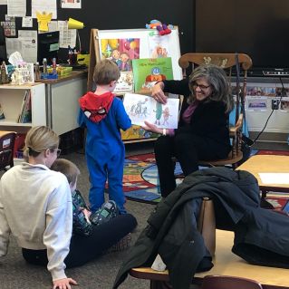 Gail reading her book to a mother and two sons, one in her lap, the other standing in front near the book.