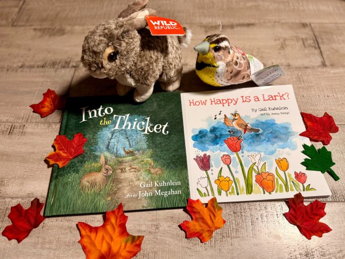 Picture books, Into the Thicket and How Happy Is a Lark? on a wood floor, a rabbit and lark stuffie and colorful leaves