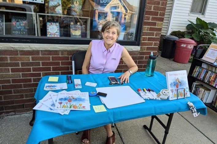 Gail sitting at a table in front of Adventure, Ink. Her book and other items on the table.