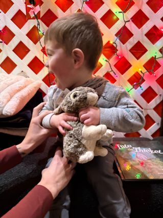 Little boy holding a rabbit stuffie and book by his side. Mom's arms holding him up on the bench.
