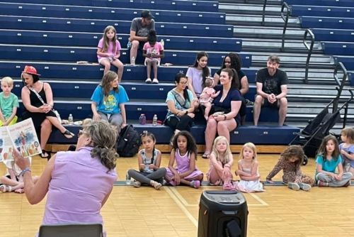Gail reading her book to children and parents in a gymnasium, children seated on the floor and some people are in the stands