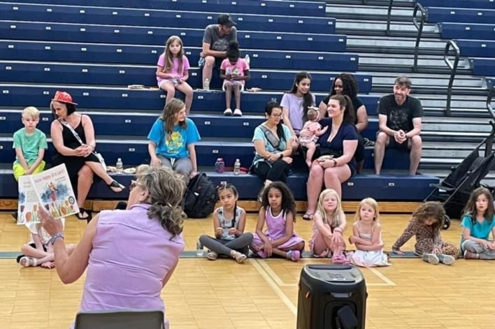 Gail reading her book to children and parents in a gymnasium, children seated on the floor and some people are in the stands