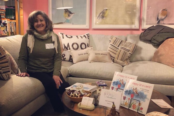 Gail sitting on a couch at the Leon and Lulu Books and Authors event with a table set up with her book on it