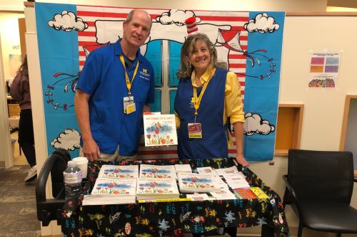 Tim and Gail in volunteer attire, with Dr Seuss backdrop and cart with How Happy Is a Lark to donate
